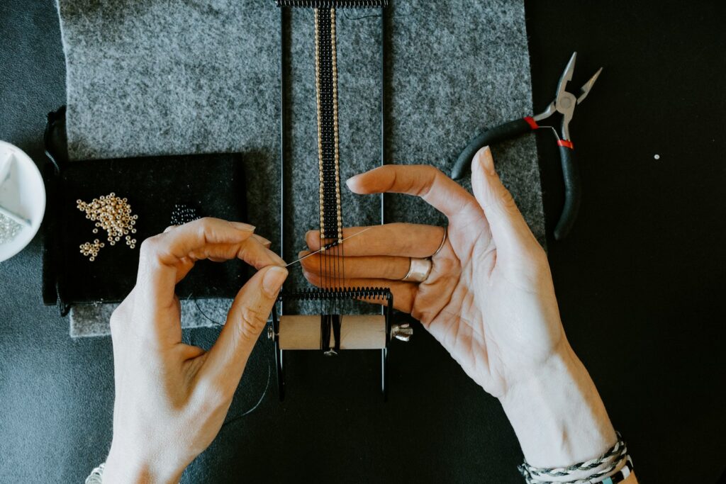 Mains d’une créatrice tissant un bracelet de perles sur un métier à tisser, pince et perles dorées posées à côté